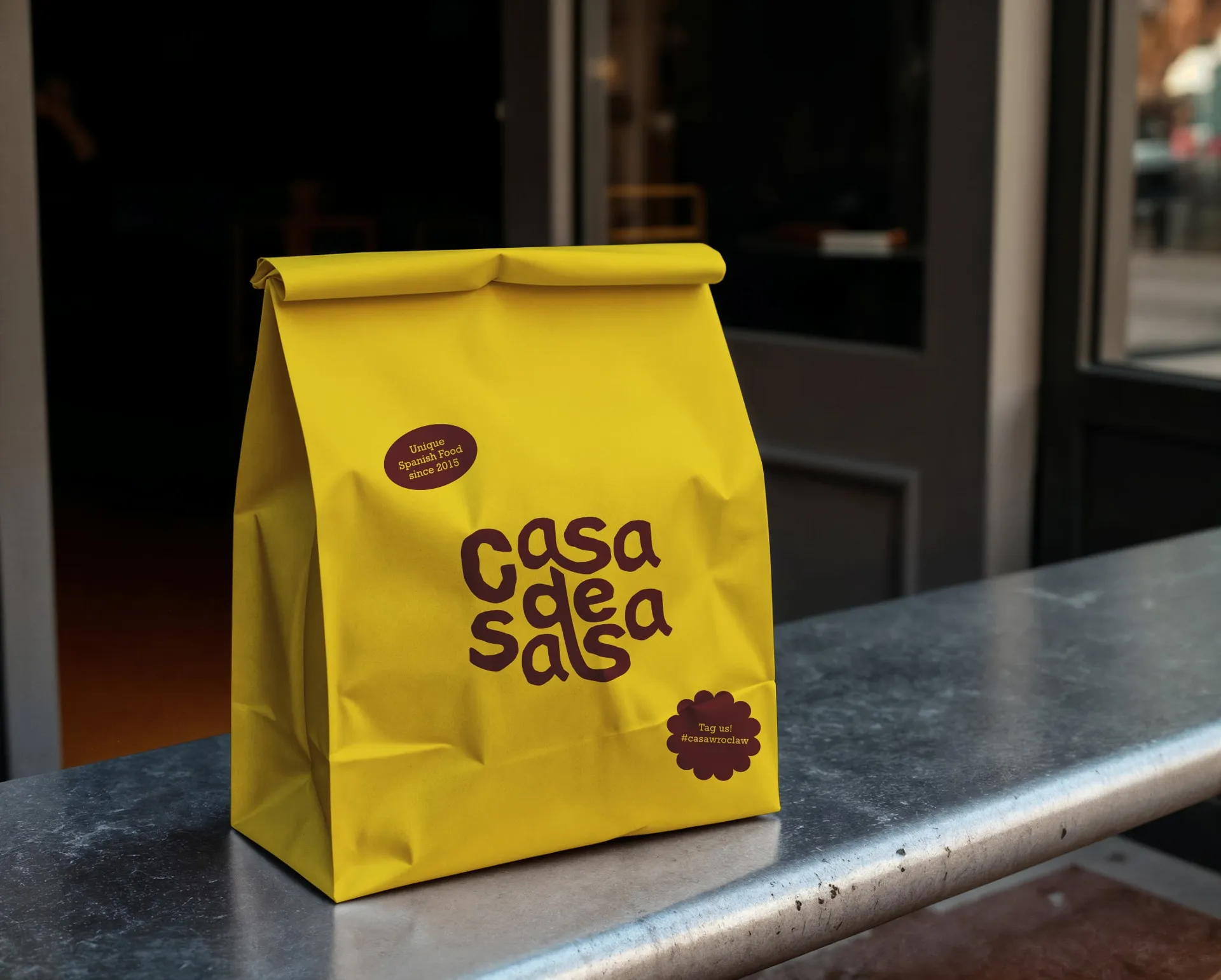 Żółta papierowa torba na wynos z napisem 'casa de salsa' i hasłem 'Unique Spanish Food since 2015', ustawiona przed wejściem do restauracji