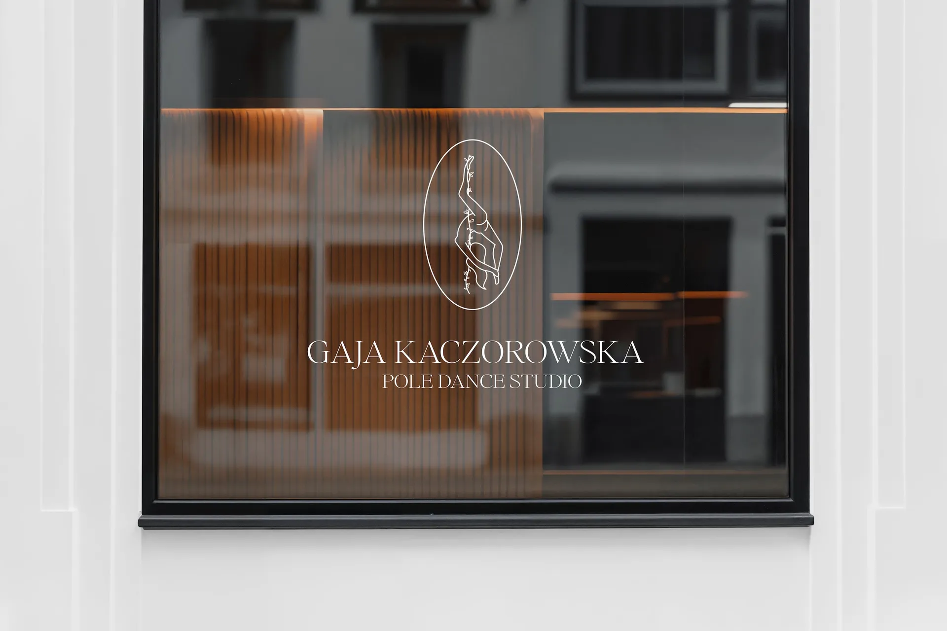 Logo Gaja Kaczorowska Pole Dance Studio na witrynie studia – biały napis i sygnatura tancerki na ciemnym szkle
